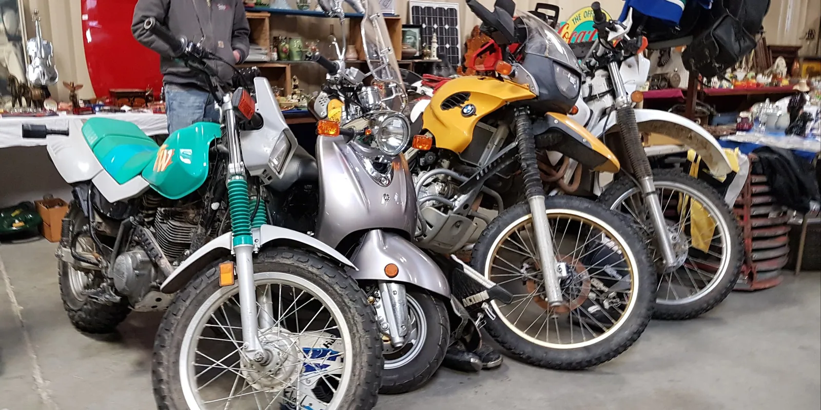 Fila de motos usadas a la venta en una feria, con una trail verde y blanca, un scooter gris y dos motos de aventura